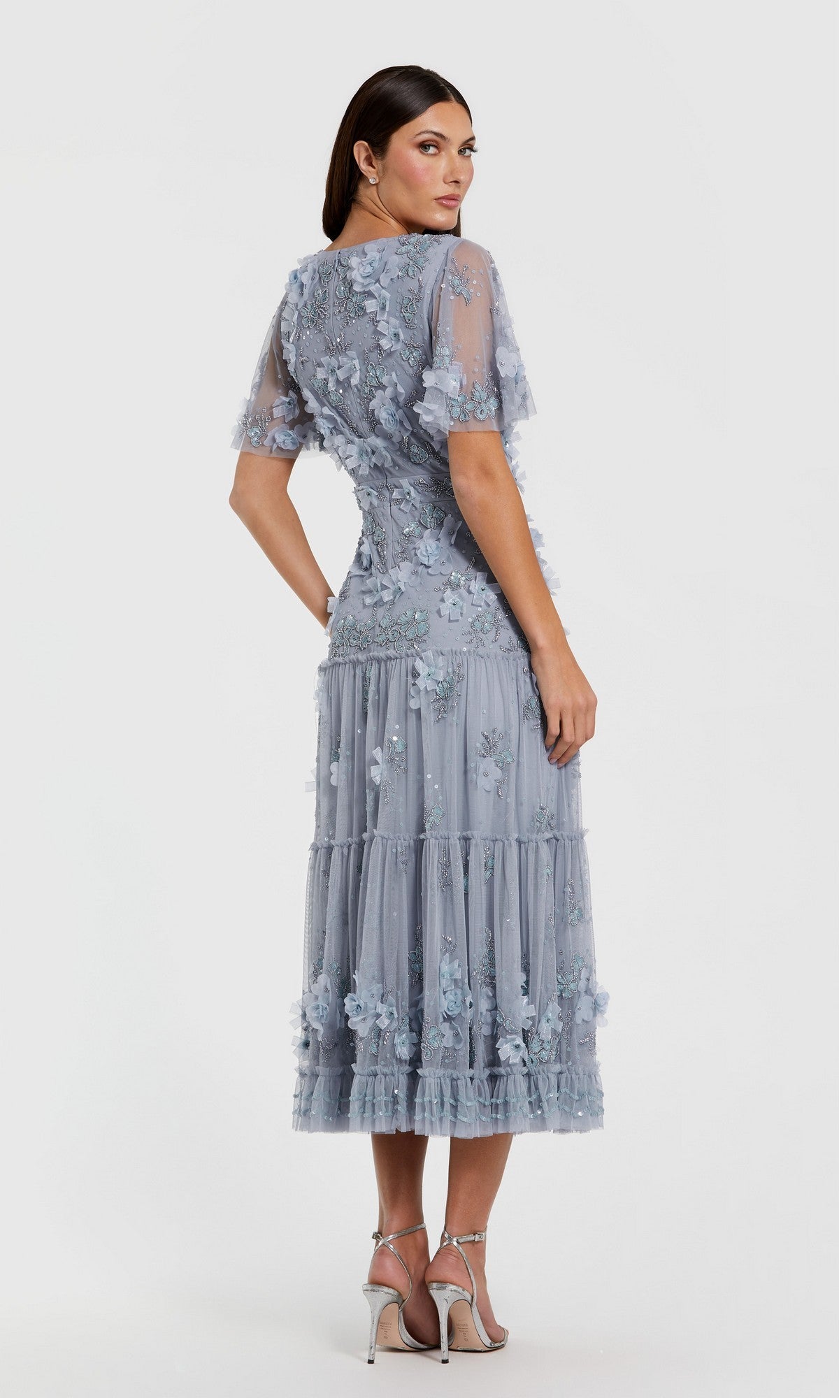 Woman wearing the Mac Duggal 9207 A-Line Wedding Guest Dress in light blue with floral embellishments, sheer short sleeves, and a tiered tea-length skirt, standing slightly turned on a plain white background.